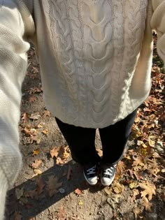 a person in white sweater and black pants standing next to leaf covered ground with their feet on the ground