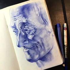 a pencil drawing of a man's face on a notebook next to two markers