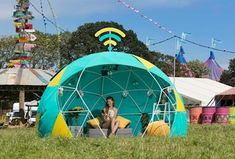 a woman sitting in a blue and yellow tent on top of a lush green field