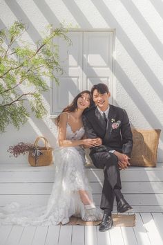 a bride and groom sitting on a bench in front of a white wall with sunlight streaming through the window