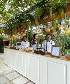 a group of people standing at a bar with plants hanging from the ceiling behind them