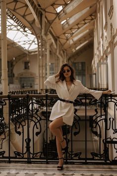 a woman standing on a balcony with her arms behind her head, wearing sunglasses and a white dress