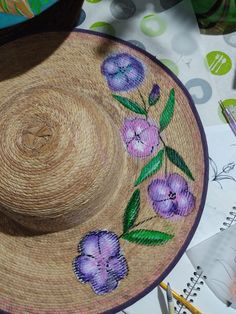 a straw hat with purple flowers painted on it and some crayons in the background