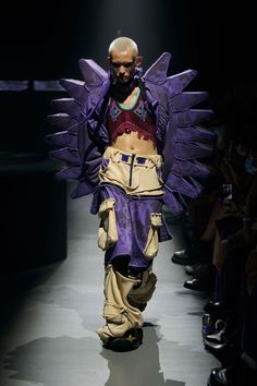 a man walking down a runway wearing purple and yellow clothing with wings on his back