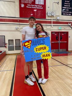 two people standing on a basketball court holding a sign that says adidas and super man