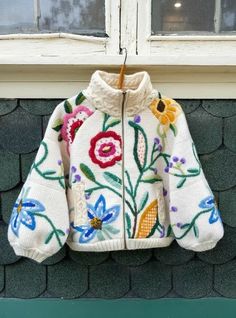 a white jacket with colorful flowers on it hanging from a window sill in front of a brick wall