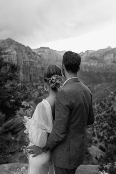 a man and woman standing next to each other in front of the mountains with wine glasses