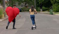 two people riding scooters down the street with a heart shaped pillow on their back