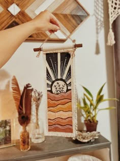 a person holding up a wall hanging with sun and tree designs on it in front of a table