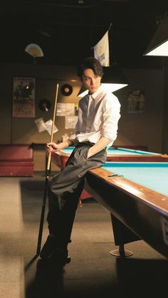 a man sitting on top of a pool table with a cue in his hand next to a pool table