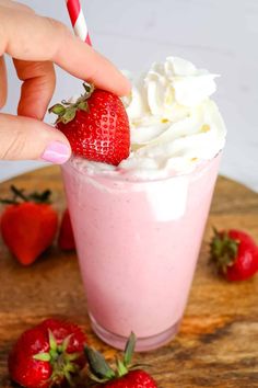 a hand is holding a strawberries milkshake with whipped cream on top and some strawberries around it