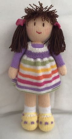 a knitted doll with brown hair wearing a striped dress and yellow shoes, sitting on a white surface