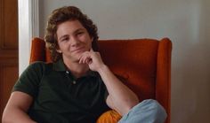 a man sitting in an orange chair with his hand on his chin and looking at the camera