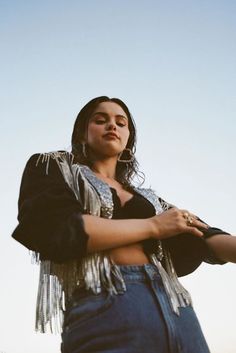 a woman standing with her arms crossed in front of the camera, wearing a fringe jacket and jeans