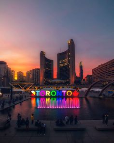 people are standing around in front of the water at night with lights on it and buildings behind them