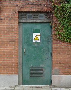 a green door with a yellow warning sign on it next to a brick wall covered in vines