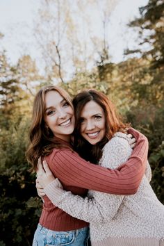 two young women hugging each other in front of some trees and bushes with their arms around one another