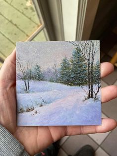 someone is holding up a small card with a snowy scene in the background and trees