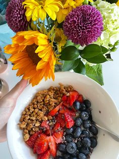 a bowl filled with granola, strawberries and blueberries next to a bouquet of flowers