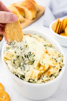 a hand dipping a piece of bread into a bowl of spinach artichoke dip