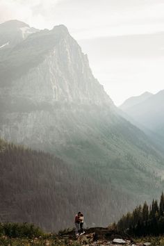 two people standing on the edge of a cliff with mountains in the backgroud