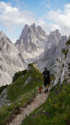 a man walking down a trail with his dog in the mountains behind him on a sunny day