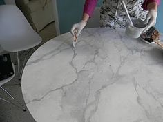 a woman in white gloves is pouring cement onto a round table with two chairs and a blue wall behind her