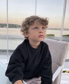 a young boy sitting on top of a chair next to a window looking out at the water