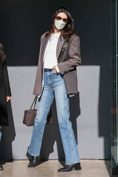 a woman wearing a face mask and jeans is walking in front of a building with her hand on her hip