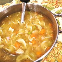 a pot filled with soup and noodles on top of a floral table cloth next to a spoon