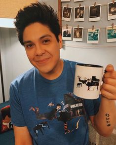 a man holding a coffee mug in his right hand and smiling at the camera with pictures on the wall behind him