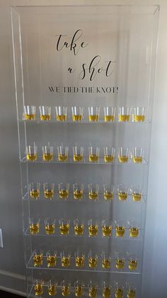 a clear display case filled with lots of glasses and gold rimmed shot glasses in front of a sign that says take a shot we tied the knot