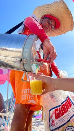 a man pouring orange juice into a cup on the beach with a straw hat over his head