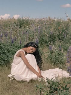 a woman in a white dress sitting on the ground next to purple flowers and grass