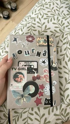 a hand holding a notebook covered in stickers and magnets on top of a bed