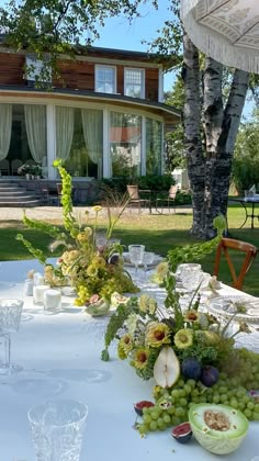 the table is set with fruit and flowers for an outdoor dinner in front of a large house