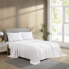 a bed with white sheets and pillows in a room next to a large window that has wooden slats on it