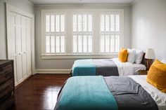 two beds in a room with wooden floors and white walls, one is blue and the other has yellow pillows