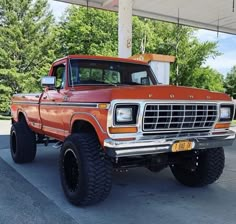 an orange pickup truck parked under a covered parking lot next to a gas station with trees in the background