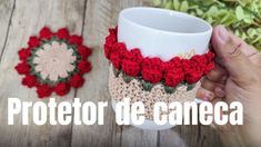 a hand holding a coffee cup with crocheted red flowers on the bottom and green leaves behind it