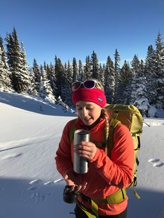 a woman with a backpack and goggles holding a cup in her hand while walking through the snow