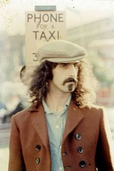 a man with long hair wearing a brown jacket and hat standing in front of a phone for a taxi sign