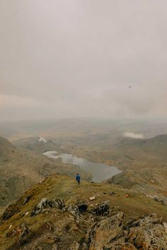 a man standing on top of a mountain next to a lake in the middle of nowhere