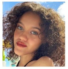 a woman with curly hair and blue eyes posing for a photo in front of the ocean