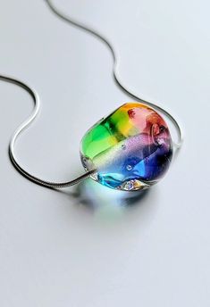 "This pendant features a rainbow hued handmade flamework glass pebble strung on a sterling silver snake chain. Each tactile 'pebble' is completely unique, built from layers of transparent colours encased in clear glass and handshaped in the torch flame. All glass is kiln annealed for durability before combining with an 18\" or 20\" sterling silver snake chain." Modern Handmade Rainbow Jewelry, Handmade Rainbow Glass Jewelry, Silver Murano Glass Round Pendant Necklace, Modern Rainbow Colored Jewelry For Gifts, Modern Rainbow Jewelry Gift, Modern Rainbow Jewelry For Gifts, Modern Rainbow Jewelry For Gift, Unique Iridescent Glass Necklaces, Rainbow Glass Jewelry Gift