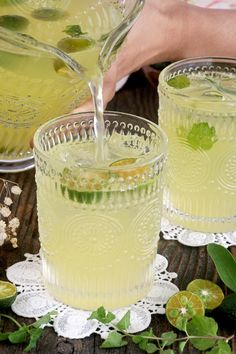 two glasses filled with lemonade and limes on top of a wooden table next to green leaves