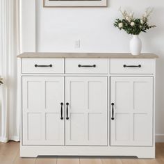 a white cabinet with two doors and some flowers in a vase next to it on top of a hard wood floor