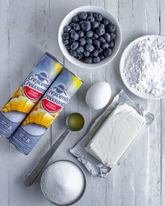 ingredients for blueberry cheesecake laid out on a white wooden table with eggs, butter and flour