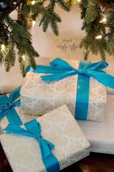 three wrapped presents under a christmas tree with blue ribbon and lights on the branches behind them