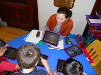 a woman sitting at a table surrounded by children using electronic gadgets and tablet computers